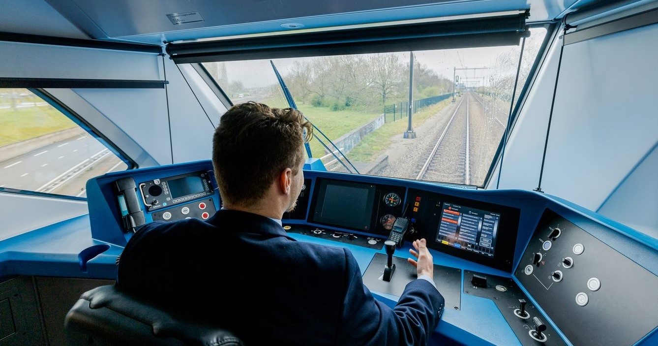 Op de afbeelding is een NS machinist te zien die een trein aan het besturen is. In de cabine zijn veel knoppen te zien en het uitzicht op het spoor.