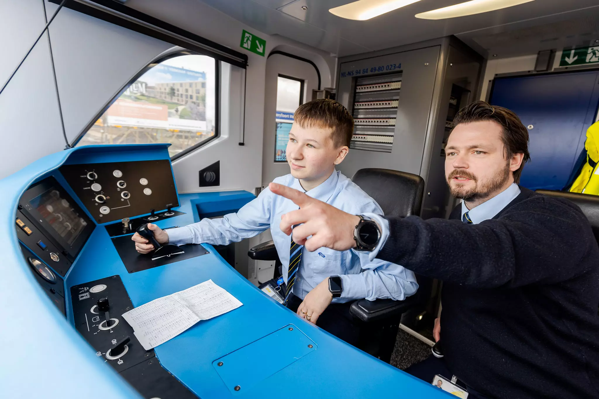 Leerling machinist krijgt instructies van een machinist in de cabine van een NS trein.